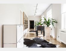 Ein heller, moderner Korridor mit großen Fenstern, weißen Wänden und glänzenden Fliesen. Darin befinden sich ein Kickertisch, große grüne Pflanzen in weißen Pflanzkästen, ein Schrank mit Metallrahmen und schwarze Kuhfell-Teppiche auf dem Boden.