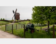 Eine historische hölzerne Windmühle steht auf einem grasbewachsenen Hügel, umgeben von Zäunen, einem Tor und einer Treppe. Im Vordergrund sind ein alter landwirtschaftlicher Anhänger und ein Baum zu sehen, eingebettet in eine ländliche Umgebung.