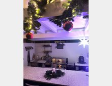 Outdoor-Küche mit einer Arbeitsplatte aus Granit, einem Edelstahlgrill, Regalen an der Wand und dekorativen Elementen. Weihnachtsdekoration mit Lichterketten, Tannenzweigen und Kugeln hängt von der Holzdecke.