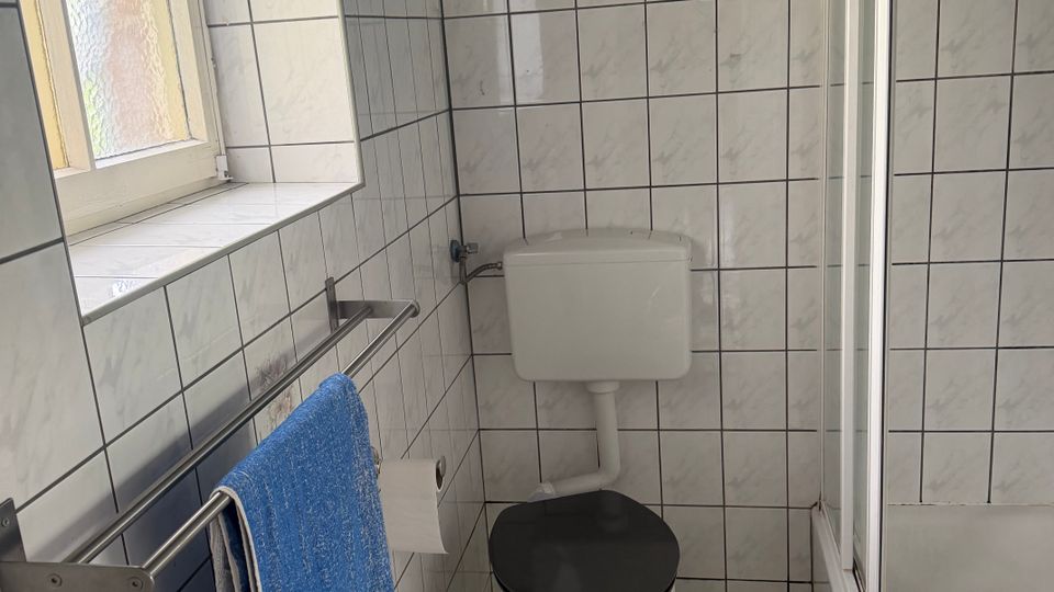 Kleines Badezimmer mit weißen, quadratischen Wand- und Bodenfliesen, Toilette mit Spülkasten an der Wand und schwarzem WC-Sitz. Links ein Milchglasfenster und ein Handtuchhalter, rechts eine Duschwanne mit Glasabtrennung.