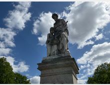 Steinskulptur auf einem rechteckigen Sockel, die zwei Personen zeigt: einen Mann mit Werkzeug und ein Kind, das einen Korb hält. Im Hintergrund ein Himmel mit weißen Wolken und umliegende Baumkronen.