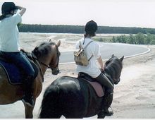 Zwei Personen reiten auf Pferden durch eine offene Landschaft mit sandigem Boden und vereinzeltem Bewuchs, im Hintergrund ist eine asphaltierte Straße und ein Wald zu sehen.
