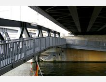 Eine Metall-Fußgängerbrücke verläuft unter einer größeren Brücke aus Stahlträgern. Darunter befindet sich ein Kanal mit ruhigem Wasser, und im Hintergrund sind Teile eines Gebäudes und eine gemauerte Wand sichtbar.