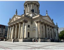 Monumentales Gebäude mit klassizistischer Architektur, flankiert von hohen Säulen und verziert mit Statuen und Reliefs im Giebel auf einer großen gepflasterten Platzfläche; im Hintergrund moderne und historische Gebäude sowie ein klarer Himmel.