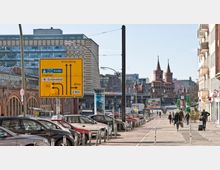 Straßenansicht in Berlin mit der Oberbaumbrücke im Hintergrund, einem markanten Backsteinbau mit zwei Türmen. Im Vordergrund befindet sich ein gelbes Verkehrsschild, das Richtungen nach Kreuzberg, Mitte und Schönefeld anzeigt, sowie parkende Autos und Fußgänger auf dem Gehweg.