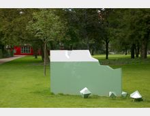 Eine moderne Skulptur mit geometrischen Formen in grüner und weißer Farbe, umgeben von einer Wiese und Bäumen in einem Park. Im Hintergrund ist ein rotes Gebäude mit großen Fenstern sichtbar.