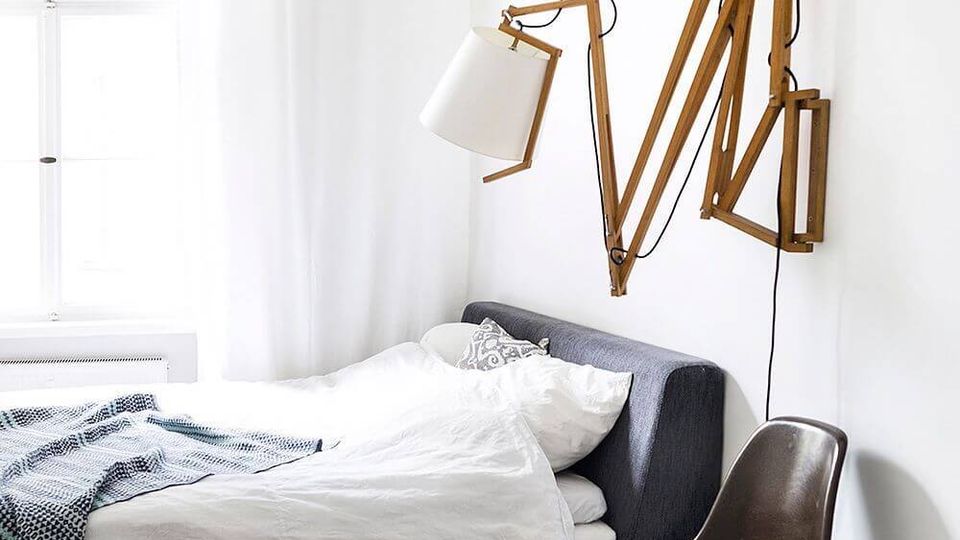 Bright minimalist bedroom with a low gray upholstered bed dressed in white bedding beside a large window with sheer white curtains. A wooden accordion wall-mounted lamp extends over the bed, and a molded chair and patterned rug sit on white-painted floorboards.