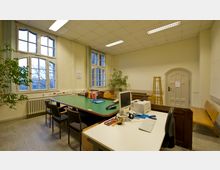 Ein heller Arbeitsraum mit hohen Decken, großen, bogenförmigen Fenstern und weißen Wänden. Der Raum enthält mehrere Tische mit Büromaterialien, einen Computer, eine Pflanze in der Ecke, eine Sitzbank an der rechten Wand und eine Holztür mit rundem Abschluss.