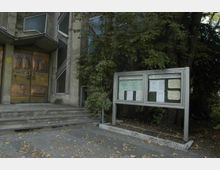 Außenansicht einer katholischen Kirche mit einem modernen Eingangsbereich aus Bronzeportalen mit Reliefs. Rechts befindet sich ein Schaukasten mit Informationen, umgeben von Bäumen und Laub auf dem Boden.