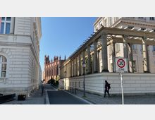 Straßenszene mit historischen Gebäuden; links ein weißes Gebäude mit Fenstern und Ornamenten, rechts ein Gebäude mit Säulen und klassizistischen Elementen. Im Hintergrund ist eine rote Backsteinkirche mit gotischen Türmen zu sehen, daneben ein Verkehrsschild mit der Aufschrift "30 Zone".