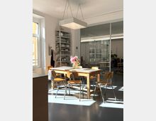 Heller Gemeinschafts- oder Pausenraum mit großem Holztisch und mehreren Stühlen, darüber eine rechteckige Hängelampe. Seitlich steht ein offenes Metallregal mit Geschirr, und im Hintergrund trennt eine Glaswand mit Türen den Raum von einem weiteren Bereich; Sonnenlicht fällt durch große Fenster auf den Boden.