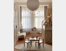 Bright children’s bedroom with large curtained windows, a round paper pendant light, and a low table with two small chairs on a circular rug. The room includes a house-frame bed with canopy curtains, a floor mattress, a rocking chair, and light wood flooring with neutral furnishings.