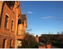 Backsteingebäude mit Spitzdach und verzierten Holzbalken, umgeben von einem Garten mit Hecken, Weg und Blumentopf im Vordergrund. Klarer blauer Himmel im Hintergrund.