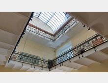 Blick nach oben in ein Treppenhaus mit einem verzierten Metallgeländer und einem Dach aus wellenförmigem Glas. Das Geländer umgibt einen rechteckigen Lichthof, und die Wände sind hellgelb gestrichen.