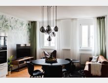 Wohnzimmer mit modernen Elementen: ein runder Esstisch mit schwarzen Stühlen, darüber eine Hängelampe mit mehreren kugelförmigen Leuchten. Im Hintergrund eine helle Wand mit Pflanzenmuster, ein Fernseher auf einem Holztisch, zwei große Fenster mit grünen und weißen Vorhängen, sowie eine braune Couch mit Decke.