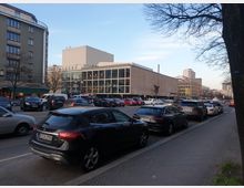 Straßenszene in einer Stadt mit parkenden Autos am Fahrbahnrand und einer stark befahrenen Straße. Im Hintergrund ein modernes Gebäude mit einer Glasfassade und einem rechteckigen Design sowie weitere mehrstöckige Gebäude, darunter ein abgerundetes Wohnhaus.