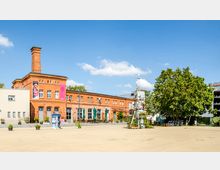 Backsteinbau mit Schornstein und großen Fenstern, links angrenzend an einen modernen, weißen Anbau. Vor dem Gebäude befindet sich ein Platz mit Sitzbereichen und Pflanzen, rechts steht ein kreativer Turm aus recycelten Materialien sowie ein großer Baum. Der Himmel ist klar und blau.