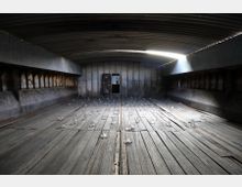 Innenraum eines rostigen, industriellen Containers mit Holzboden, auf dem mehrere kleine durchsichtige Glaselemente verteilt sind. Im Hintergrund befindet sich eine Metalltür mit einem kleinen Fenster, in dem das Gesicht einer Person abgebildet ist.