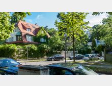 Straßenszene in einer grünen Wohngegend mit einem großen, von Efeu bewachsenen Haus mit roten Dachziegeln und Fenstern mit weißen Rahmen im Hintergrund. Im Vordergrund stehen Autos geparkt entlang einer Kopfsteinpflasterstraße, umgeben von Bäumen und einem Straßenschild mit den Namen „Niklasstraße“ und „Kaunstraße“.