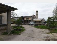 Ein Industriegelände mit mehreren flachen Gebäuden aus Beton und Ziegeln, darunter ein Atelier mit ausgeschildertem Eingang. Im Hinterhof stehen ein Transporter, Holzpaletten und vereinzelte Pflanzen, umgeben von einer Betonfläche und vereinzeltem Grün. Im Hintergrund ist ein hoher Schornstein zu sehen.