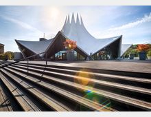 Das Bild zeigt das berühmte Tempodrom in Berlin, ein modernes Veranstaltungsgebäude mit einem auffälligen, weißen, zeltartigen Dach aus Stahl und Beton. Im Vordergrund befinden sich breite Holzstufen, während bunte Herbstbäume und große Pflanzenkübel die Umgebung schmücken.