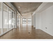 Leerer, moderner Büroraum mit hellem Holzboden, großen Glaswänden auf der linken Seite und sichtbarem Beton an der Decke. Im Hintergrund sind bodentiefe Fenster mit viel Tageslichteinfall zu sehen.