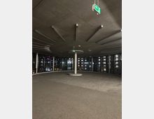 Ein leerer, runder Innenraum mit Betonboden und -decke, durchzogen von schmalen Deckenverstrebungen. Der Raum ist von bodentiefen Fenstern umgeben, durch die Gebäude bei Nacht sichtbar sind. Über einem zentralen Pfeiler hängt ein grünes Notausgangsschild.