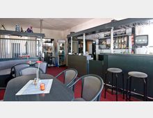 Ein kleiner Bistro- oder Cafébereich mit roten Teppichboden, schwarzen Tischen und grauen Stühlen. Im Hintergrund eine Theke mit Glasregalen, auf denen verschiedene Flaschen stehen, daneben ein Kühlschrank, sowie eine kleine Sitzbank und ein Fernseher an der Wand.