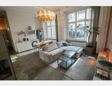 Wohnbereich einer modernen Loftwohnung mit hohen Decken, großen Fenstern mit Gitter, weiß gestrichenen Ziegelwänden und Betonboden. Der Raum ist mit einer offenen Küche, einer grauen Eckcouch, einem Esstisch mit Holzstühlen, einem großen Topfpflanzen und einer Deckenlampe mit Glasdetails ausgestattet.