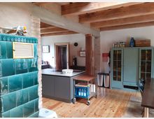 Rustikale Wohnküche mit sichtbaren Holzbalken, Holzfußboden und gemauerten Stützen. Links ein grüner Kachelofen, in der Mitte eine Kücheninsel mit Kochfeld und rechts ein großer Vitrinenschrank sowie ein Esstischbereich.