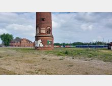 Ein altes, rundes Backsteingebäude mit Fenstern und Graffiti steht auf einer offenen, teils bewachsenen Fläche. Im Hintergrund sind ein historisches Backsteingebäude sowie mehrere bunte Züge und Bäume sichtbar.