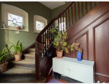 Treppenhaus mit geschwungener Holztreppe und dunklem Geländer, flankiert von zwei kleinen Fenstern mit dekorativem Gitter. Im Eingangsbereich stehen mehrere Topfpflanzen und ein weißes Sideboard vor einer holzvertäfelten Wand.