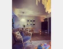 Wohnzimmer mit modernem, skandinavisch inspiriertem Design. Im Vordergrund ein Sessel aus Rattan mit blauen Kissen, ein Holz-Couchtisch mit Büchern und einer Vase, im Hintergrund ein Esstisch mit Stühlen, eine Glaspendelleuchte und mehrere gerahmte Schwarz-Weiß-Bilder an der Wand.