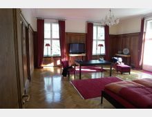 Ein elegantes Wohnzimmer mit Holzwandverkleidungen, großen Fenstern mit roten Vorhängen und einem glänzenden Holzparkettboden. Der Raum ist mit einem Tisch, einem Sofa, einem Fernseher und einem Kronleuchter ausgestattet, dazu passend rote Teppiche und Sitzmöbel.