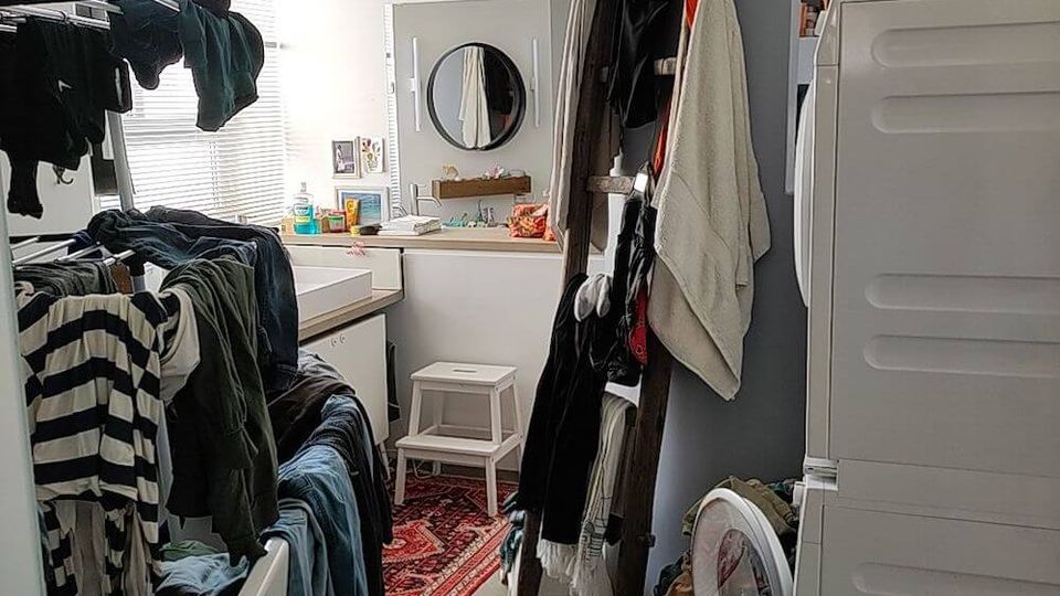 A cluttered laundry room with a tiled floor, featuring a washing machine, stacked dryer, and drying racks filled with clothes. The space also includes a curved wall, a small sink with a circular mirror, and various household items scattered throughout.