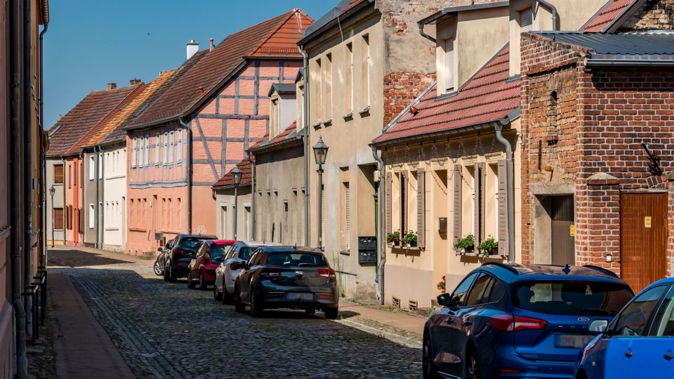 Blick entlang einer schmalen Kopfsteinpflasterstraße in einem Altstadtviertel, gesäumt von dicht stehenden Wohnhäusern mit Putzfassaden und teils sichtbarem Fachwerk sowie Ziegelmauern. Am Straßenrand parken mehrere Autos; Laternen und rot gedeckte Satteldächer prägen das Straßenbild.