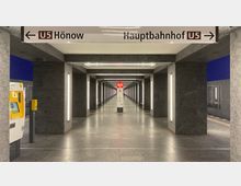 U-Bahn-Station mit einem hell beleuchteten Gang aus grauem Stein, flankiert von Säulen mit vertikalen Lichtleisten. Oben hängen Wegweiser mit der Beschriftung „U5 Hönow“ und „Hauptbahnhof“, im Hintergrund steht eine SOS-Säule.