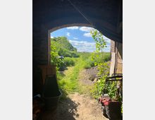 Blick durch eine steinerne Toröffnung auf einen grasbewachsenen Pfad, der durch eine grüne Landschaft führt. Im Vordergrund sind landwirtschaftliche Geräte zu sehen, darunter ein Rasenmäher, während im Hintergrund Bäume und blauer Himmel mit weißen Wolken sichtbar sind.