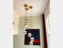 Moderne Hängeleuchte mit mehreren kugelförmigen, weißen Lampenschirmen, die an dünnen Kabeln in einer geschwungenen Anordnung von goldenen Deckenhalterungen hängen. Im Hintergrund ist eine hellgrüne Wand mit einem Gemälde in dunklen Farben und roten Akzenten zu sehen, daneben ein Fenster mit einem dunkelroten Vorhang.