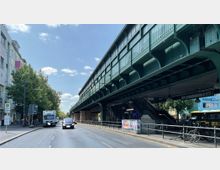Eine Straßenszene mit einer erhöhten U-Bahn-Strecke auf grüner Stahlkonstruktion, darunter eine Treppe zu einem Bahnsteig. Links stehen Gebäude und Bäume, rechts parken Fahrräder und ein Bus ist sichtbar.