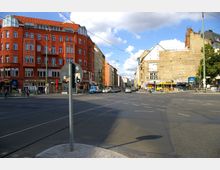 Straßenkreuzung in einer Stadt mit Schienen für Straßenbahnen auf der Fahrbahn. Links steht ein mehrstöckiges, rotes Gebäude mit Geschäften im Erdgeschoss, rechts sind weitere Gebäude, teils mit sichtbarer Ziegelwand und Fassadenbeschriftungen. Im Hintergrund sind Autos und Fußgänger zu sehen, sowie ein U-Bahn-Zugang.