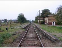 Ehemaliger ländlicher Bahnhof mit einem Gleis in der Mitte, flankiert von verfallenen Bahnsteigen und einem gelben Backsteingebäude. Die Umgebung besteht aus Wiesen, vereinzelten Bäumen und vereinzelten Häusern im Hintergrund.