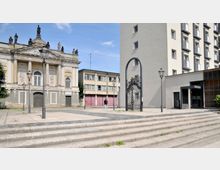 Barockgebäude mit Säulen und Figuren auf dem Gesims links, flankiert von moderneren Gebäuden im Hintergrund und rechts. Im Vordergrund gepflasterter Platz mit Treppenstufen und schmiedeeisernes Tor.