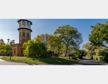 Backsteinturm in einem Park mit umliegenden Bäumen, Rasenflächen und einem asphaltierten Weg. Der Turm hat eine achteckige, verglaste Spitze und steht in sonniger Umgebung mit vereinzelten Straßenlaternen.