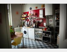 Kleine Küche mit rot gefliester Wand, offenen Regalen voller Kochutensilien, einem Herd, einer Waschmaschine und einer Spülmaschine. Der Boden ist in einem Schachbrettmuster aus schwarzen und weißen Fliesen gestaltet, mit einem kleinen runden Holztisch und gelbem Stuhl im Vordergrund.