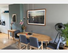 Essbereich einer Wohnung mit einem ovalen Holztisch, umgeben von vier dunkelblauen Polsterstühlen. An der grauen Wand hängt ein großes gerahmtes Bild, daneben stehen eine Vase mit Blumen und eine große Stehleuchte; der Boden ist mit Parkett ausgelegt.