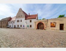 Historisches Gebäude mit weißer Fassade und rotem Satteldach, gekennzeichnet durch einen verzierten Stufengiebel, neben einer Mauer mit großem Holztor, das von einem roten Ziegelbogen gerahmt ist. Im Vordergrund liegt eine gepflasterte Straße, und an der Mauer sind Informationsplakate und eine Wanddekoration sichtbar.