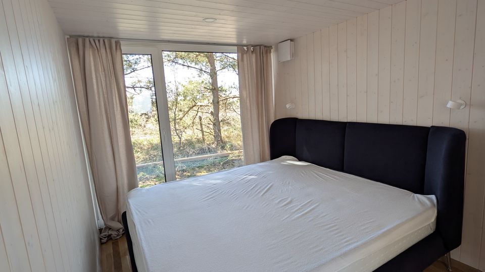 Schmales Schlafzimmer mit hellen Holzpaneelen an Wänden und Decke, Holzboden und einem großen Bett mit dunklem Polsterkopfteil. Am Ende des Raums befindet sich eine bodentiefe Glasschiebetür mit Vorhängen und Blick ins Grüne.