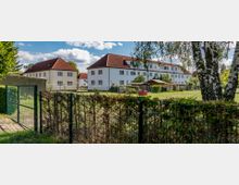 Mehrfamilienhäuser mit weißen Fassaden und roten Satteldächern stehen in einer grünen Wohnsiedlung. Im Vordergrund sind ein Zaun, Büsche, Bäume sowie Rasenflächen zu sehen.