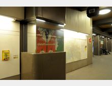 U-Bahn-Station mit beleuchteten Wänden und einer großen Wandkarte. Im Vordergrund befindet sich die Beschilderung „Mehringdammplatz“ auf einer bunten Fliesenwand, während im Hintergrund Sitzbänke unter Deckenleuchten sichtbar sind.